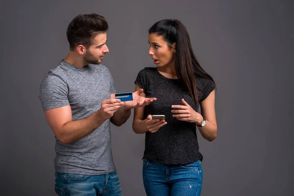 双方の思いとスマート フォンを使用している彼の美しいガール フレンドに彼のクレジット カードを示すハンサムな若い男を混乱させる — ストック写真