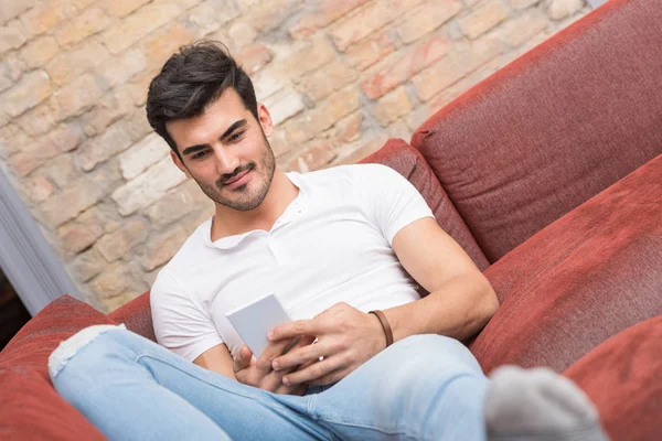 Um jovem feliz deitado em um sofá e usando seu smartphone — Fotografia de Stock