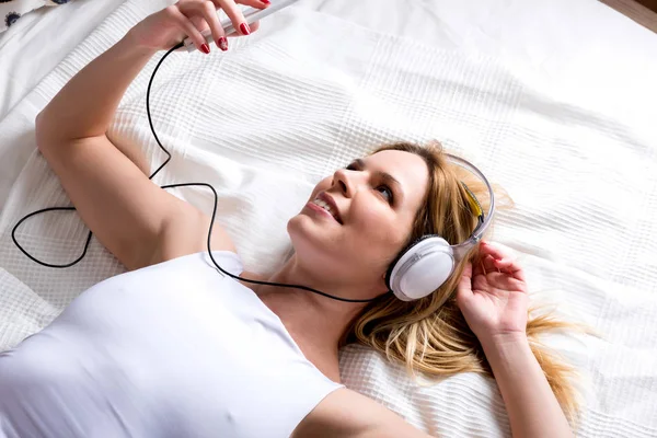 Een lachende jonge vrouw liggend op een bed en luisteren naar muziek — Stockfoto