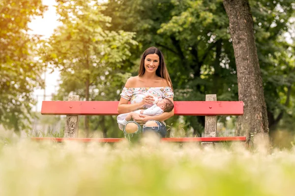 Una joven madre sentada en un banco y alimentando a su bebé — Foto de Stock