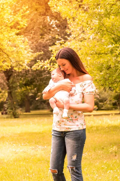 Una joven madre de pie en el parque y sosteniendo a su bebé — Foto de Stock