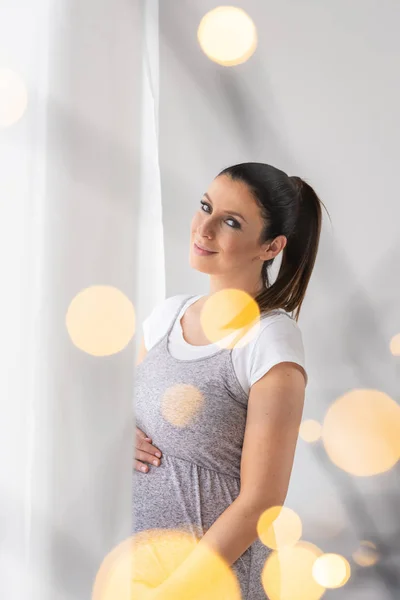 Een zwangere vrouw staat naast het venster in een elegante grijs — Stockfoto