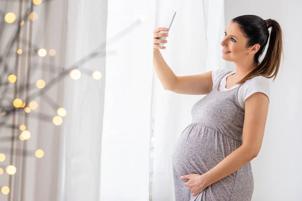 Een zwangere vrouw staat naast het venster en het nemen van een selfie — Stockfoto