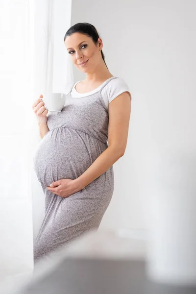 Een zwangere vrouw staat naast het venster en het drinken van koffie — Stockfoto