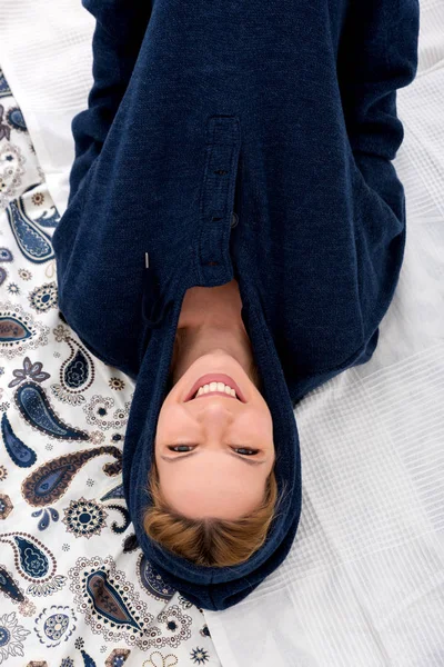 Een lachende jonge vrouw liggend op een bed in een blauwe trui — Stockfoto