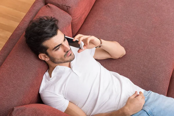 Серьезный молодой человек лежит на диване и разговаривает по телефону — стоковое фото