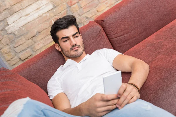 Un joven serio acostado en un sofá y usando su teléfono inteligente — Foto de Stock
