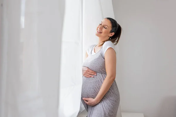 Een zwangere vrouw die naast het raam staat met handen op haar b — Stockfoto