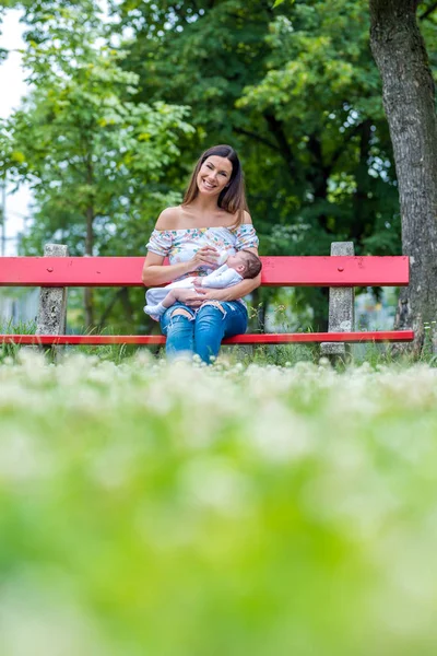 Una joven madre sentada en un banco y alimentando a su bebé — Foto de Stock