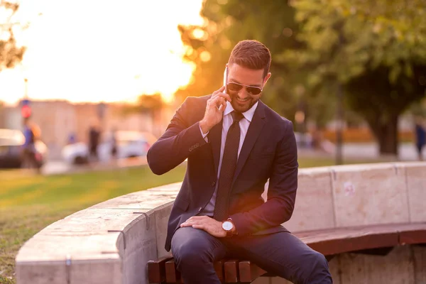 Un hombre de negocios sentado en un banco y hablando por teléfono — Foto de Stock