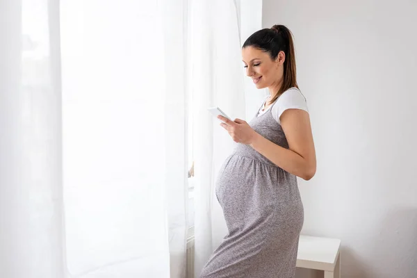 Een zwangere vrouw die naast het raam staat en haar slimme — Stockfoto