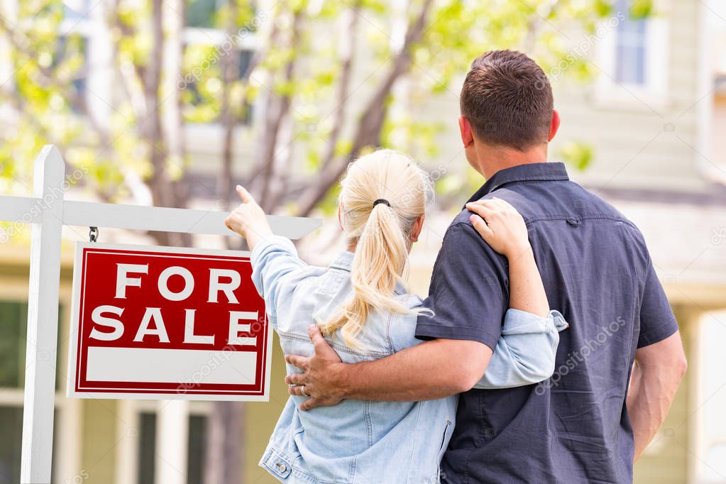Caucasian Couple Facing and Pointing to Front of For Sale Real Estate Sign and House.