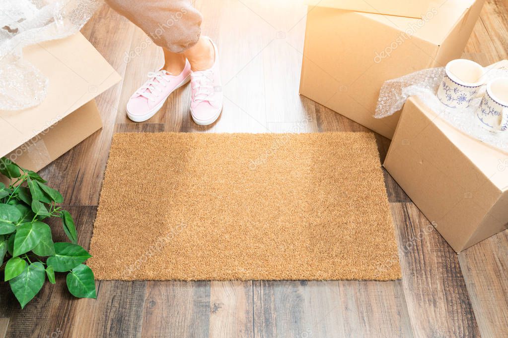 Woman in Pink Shoes and Sweats Standing Near Home Sweet Home Welcome Mat, Boxes and Plant.