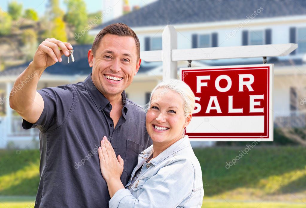 Young Adult Couple With House Keys In Front of Home and For Sale Real Estate Sign.