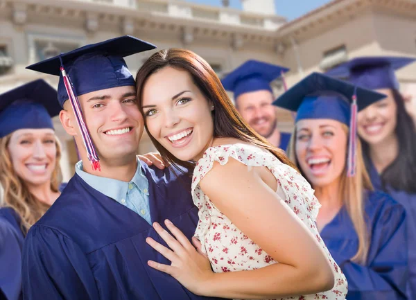 Гордый мужчина выпускник в шапке и платье с девушкой среди других выпускников позади — стоковое фото
