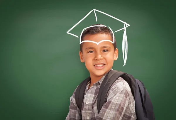 Jovem estudante hispânico menino vestindo mochila frente de quadro negro com tampa de graduação desenhado em giz sobre cabeça — Fotografia de Stock