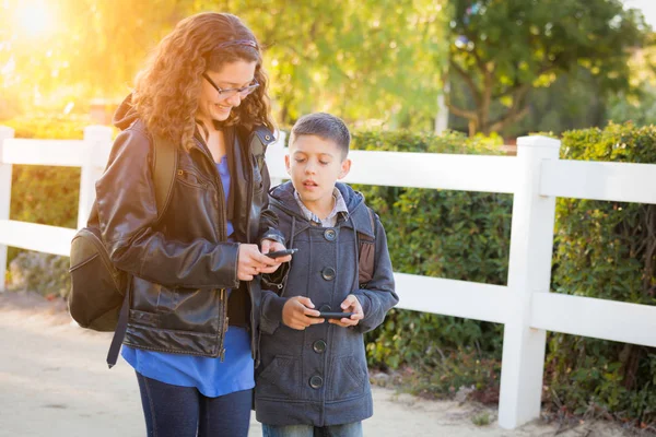 Hermano y hermana hispanos usando mochilas caminando mensajes de texto en teléfonos celulares — Foto de Stock