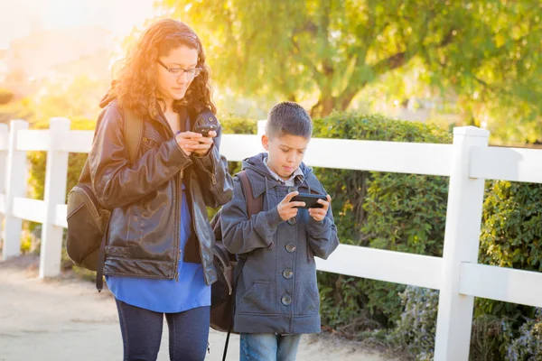Hermano y hermana hispanos usando mochilas caminando mensajes de texto en teléfonos celulares — Foto de Stock