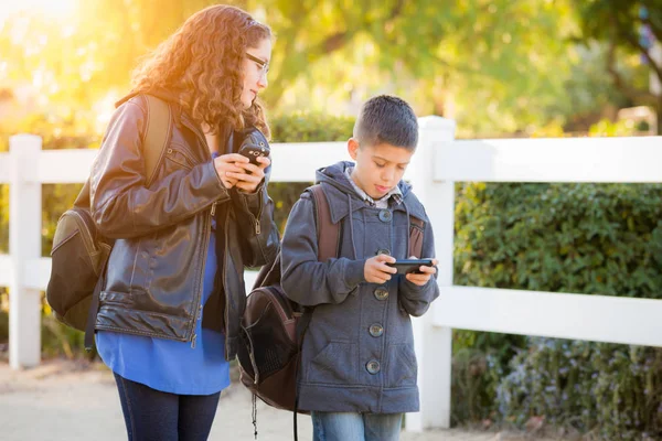 Hermano y hermana hispanos usando mochilas caminando mensajes de texto en teléfonos celulares —  Fotos de Stock