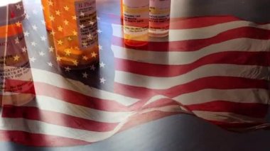 4k Slow Motion İlaç Şişeleri ve Hayalet Amerikan Bayrağı Dalgasıyla Düşen Haplar. İlaç şişesi etiketleri hayali isimler, adresler ve stok fotoğrafçılığı endüstrisi için özel olarak tasarlanmış bilgilerle tescilli değildir..