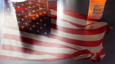 4k Slow Motion İlaç Şişeleri ve Hayalet Amerikan Bayrağı Dalgasıyla Düşen Haplar. İlaç şişesi etiketleri hayali isimler, adresler ve stok fotoğrafçılığı endüstrisi için özel olarak tasarlanmış bilgilerle tescilli değildir..