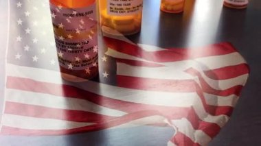 4k Slow Motion İlaç Şişeleri ve Hayalet Amerikan Bayrağı Dalgasıyla Düşen Haplar. İlaç şişesi etiketleri hayali isimler, adresler ve stok fotoğrafçılığı endüstrisi için özel olarak tasarlanmış bilgilerle tescilli değildir..