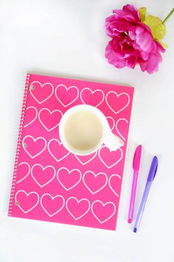 Pembe ve beyaz kalp tasarımı ile Spiral Defter. Fincan çay beyaz kupa, pembe ipek çiçek ve düz lay içinde beyaz zemin üzerine pembe ve mor kalem