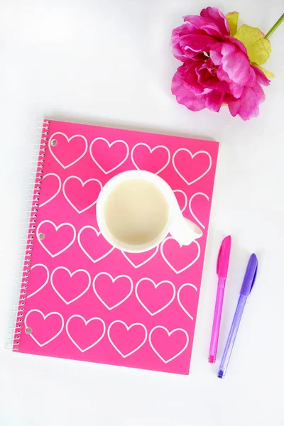 Spiral notebook with pink and white hearts design. Cup of tea in white mug, pink silk flower and pink and purple pens on white background in flat lay