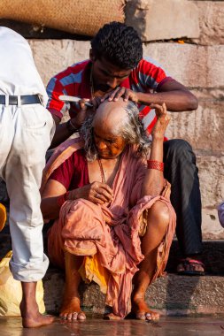 Ganj Nehri 'nin kutsal sularının yanında yaşlı kadının kafasını tıraş eden adam. Varanasi, Hindistan