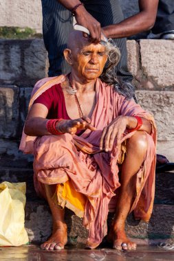 Genç adam yaşlı kadının kafasını tıraş ediyor ve Ganj Nehri 'nin kutsal sularının yanında oturuyor. Varanasi, Hindistan