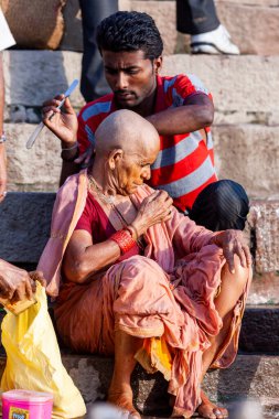 Genç adam yaşlı kadının kafasını tıraş ediyor ve Ganj Nehri 'nin kutsal sularının yanında oturuyor. Varanasi, Hindistan