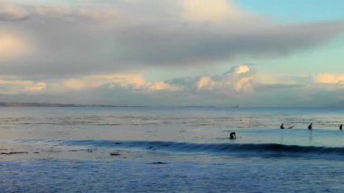 Dalgaları yakalamaya çalışan sörfçülerle birlikte denizin üzerinde muhteşem bulutlu bir gün batımı. 