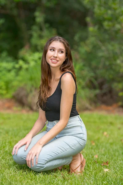 Attractive woman squatting in a park scene wearing tank top and — Stock Photo, Image