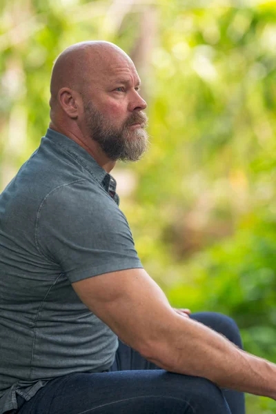 Imagem de um homem careca com barba sentada do lado de fora e olhando para longe — Fotografia de Stock