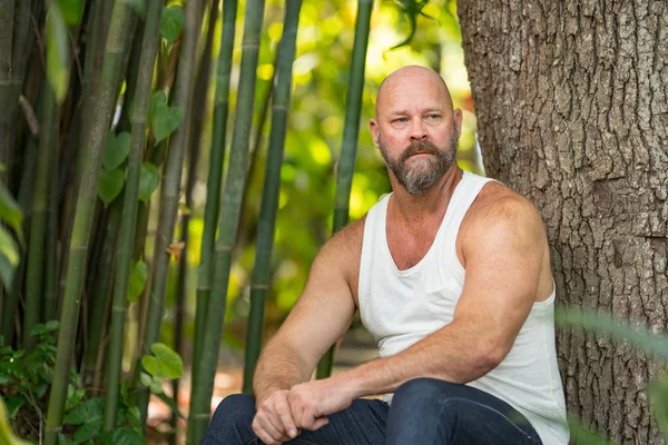 Bonito homem de tshirt sentado ao lado de uma árvore no parque — Fotografia de Stock