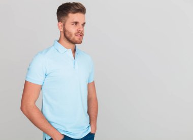 hafif mavi polo yan elleri ceplerinde ışık gri arka plan üzerinde dururken t-shirt isteyen giyen rahat adam portresi