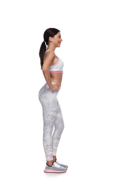 вид сбоку на сексуальную брюнетку гимнастку, держащую бедра, стоя на белом фоне. На ней пара белых леггинсов и белый топ.
