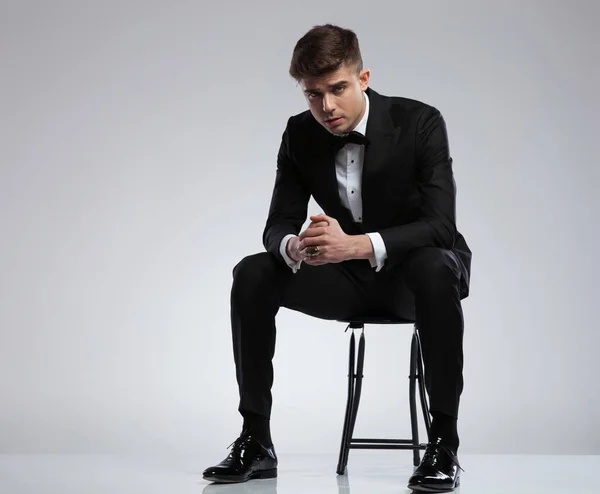 性感的年轻人穿着黑色燕尾服坐在椅子上浅灰色背景 — 图库照片
