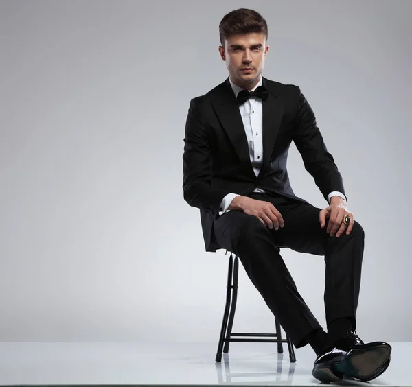 有魅力的年轻人穿着黑色燕尾服坐在金属椅上 腿交叉在浅灰色背景上 — 图库照片