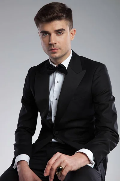 黒のネクタイと指に金の指輪を持っていること 明るい灰色の背景の上に座って黒のタキシードで魅力的な若い男の肖像 — ストック写真