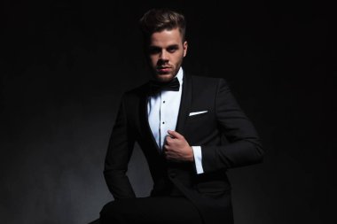 siyah arka plan oturan ve takım elbise yaka tutarak iken poz siyah smokin giymiş yakışıklı adam