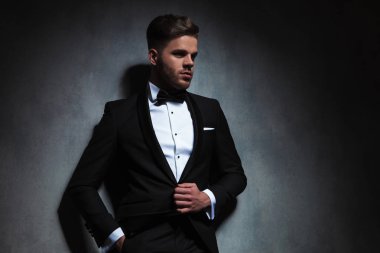 siyah smokin takım elbise yaka tutarken koyu gri duvara yaslanmış iken yan isteyen rahat şık adam portresi