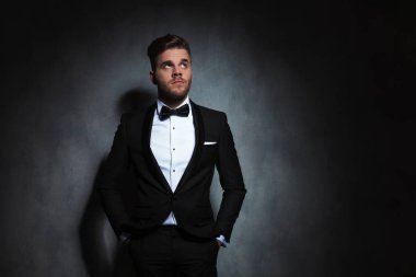 elleri ceplerinde duran zarif genç adam yan yakınındaki bir koyu gri duvar kağıdı için arama yapar. Dalgın seyir siyah bir smokin giyiyor