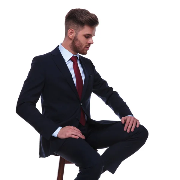 坐在椅子上的英俊的商人在白色的背景上向下看 一边把手放在大腿上 他穿着一件海军西装和一条红领带 — 图库照片