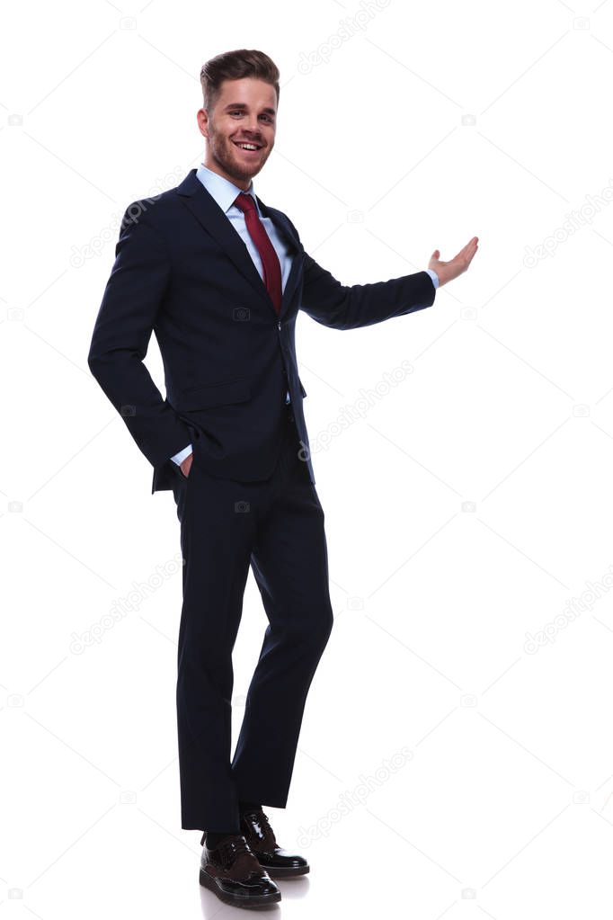 happy businessman standing with hands in pockets on white background while wearing a navy colored suit and a red tie. He is presenting with his hand