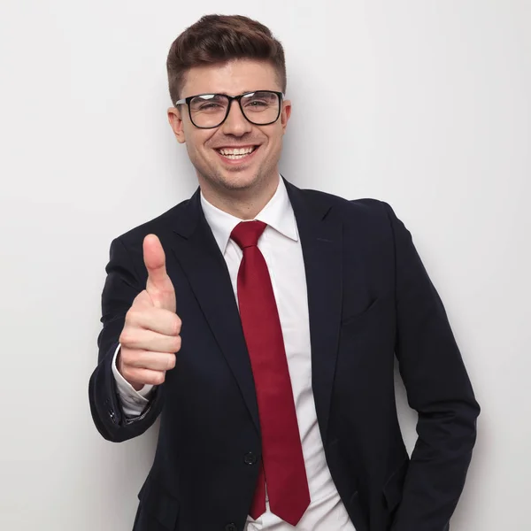 ライトグレーの背景に立っている間登録して親指を作ってうれしそうな青年実業家の肖像画 彼はネイビーのスーツ 赤いネクタイ メガネを身に着けています — ストック写真