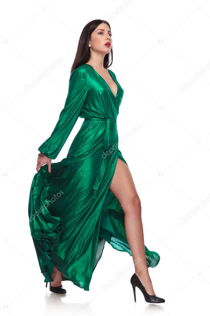 sensual woman in fluttering long green dress walks to side while holding it, on white background, full body picture