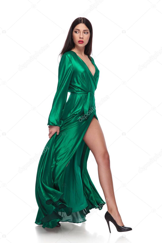 side view of woman in green dress walking on white background and looking behind, while holding the dress, full body picture