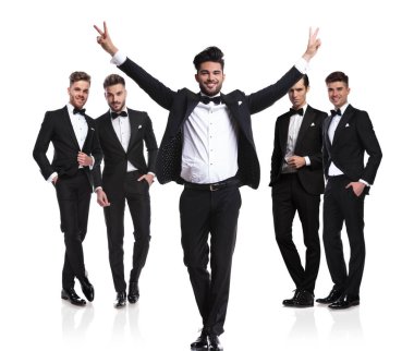 Beyaz arka planda siyah smokin giymiş, zafer işareti önünde dururken ekibi damat yapar