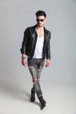 yakışıklı erkek deri ceket ve güneş gözlüğü takmış açık gri renkli cross-legged üzerinde duruyor ve tarafına görünüyor